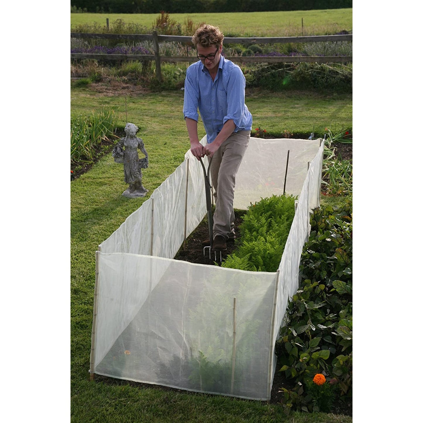 Man gardening with a sturdy micromesh pest and wind barrier enclosing a raised bed, protecting crops from pests while allowing sunlight and air in, set in a lush garden.