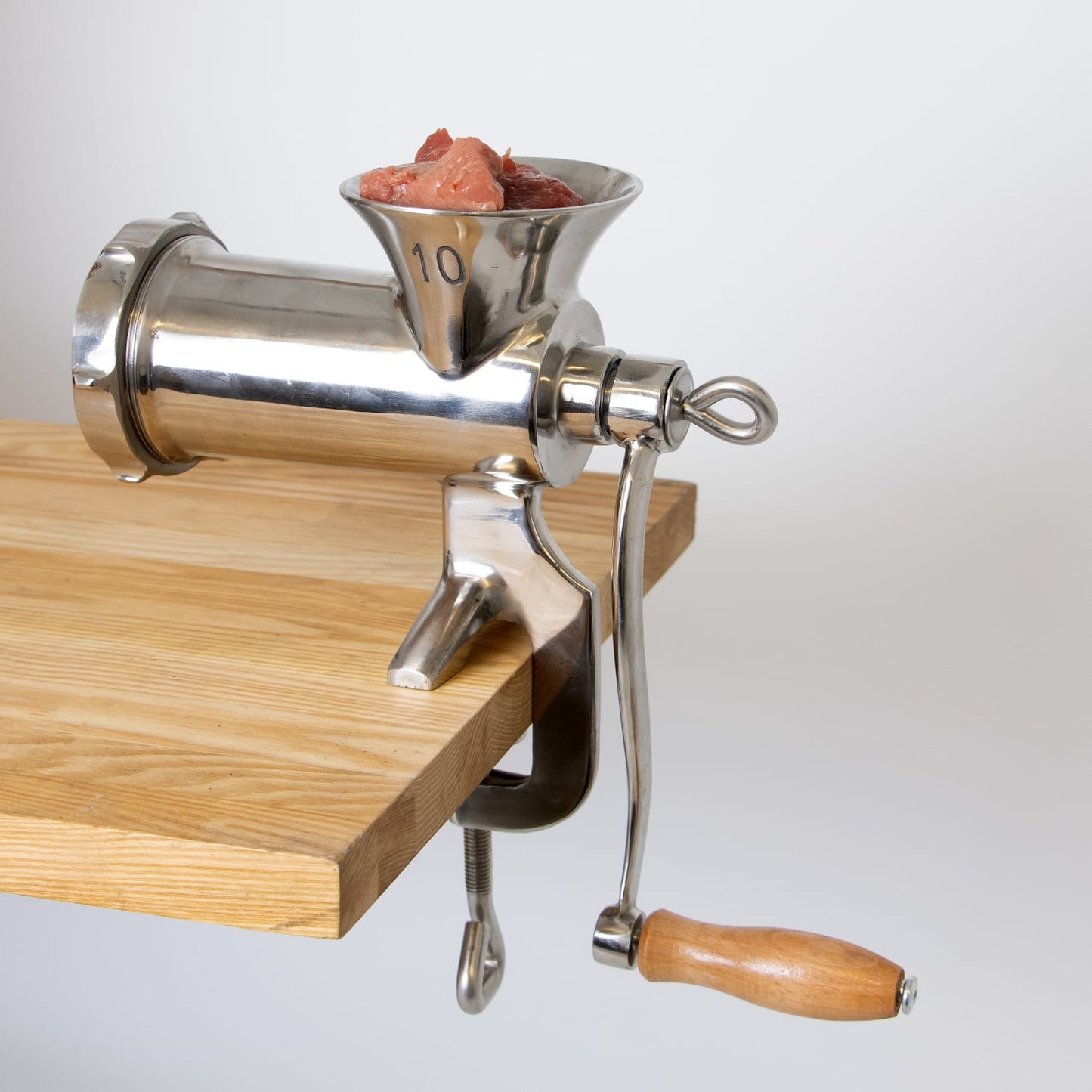 Handcrafted stainless steel clamp-down meat grinder with wooden handle secured to a wooden table, ready for fresh meat grinding, suitable for Kitchen & Food and Specialty Equipment collections.