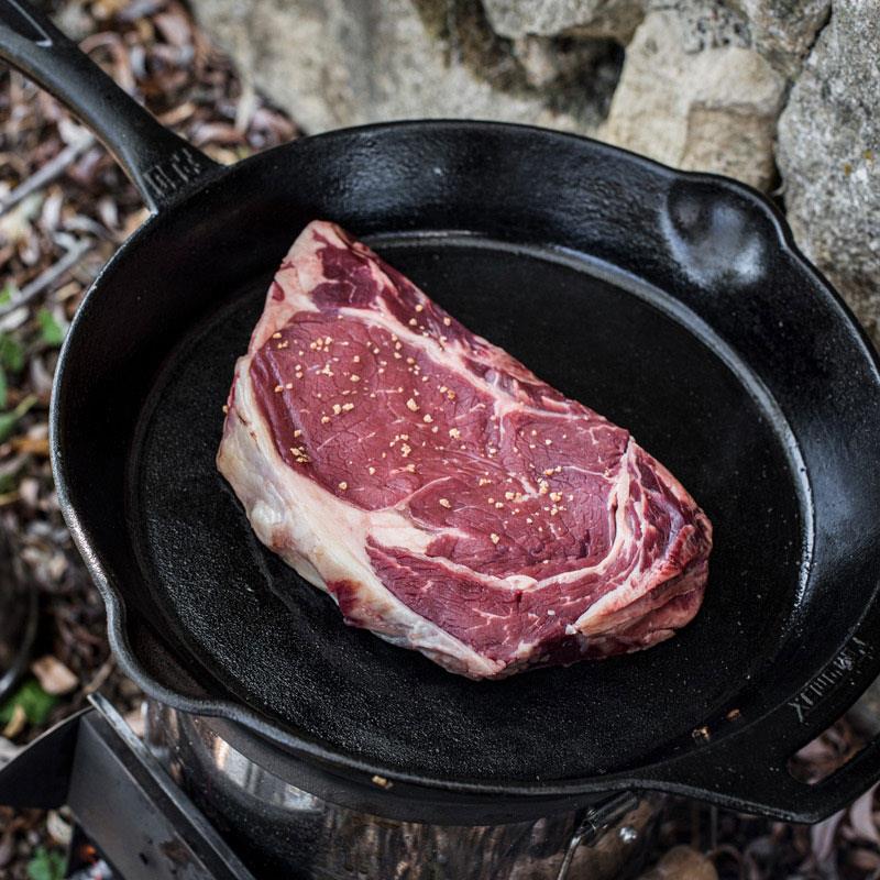 Premium cast iron skillet with long handle cooking a seasoned raw ribeye steak outdoors, perfect for Farm, Garden & Tool, Kitchen & Food, Camping, Cookware, Cast Iron, Best Sellers, and Promo Eligible collections.
