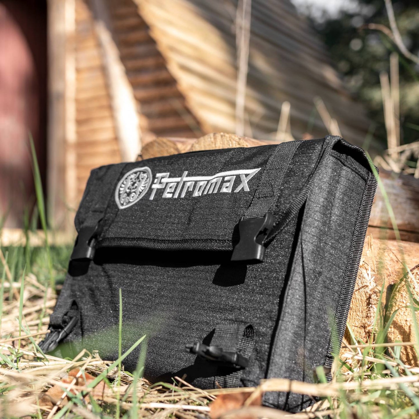 Compact black carrying case with embroidered logo, designed to store a foldable outdoor camp stove, placed on grass near stacked firewood, suited for Farm, Garden & Tool, Camping, Emergency Supplies, Best Sellers, and Promo Eligible collections.