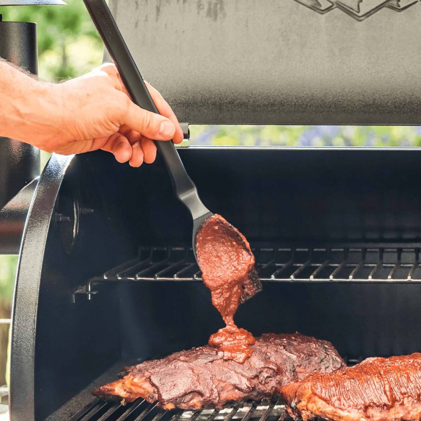 Hand using a BBQ Brizzle Basting Brush with a deep scooping head to apply barbecue sauce over ribs on a charcoal grill, perfect for Kitchen & Food, Grilling, and Kitchen Gadgets collections.