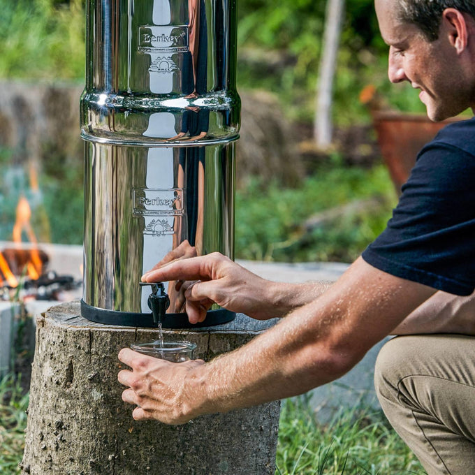 Polished stainless steel water purifier with black filter elements dispensing water into a clear glass, placed outdoors on a tree stump, ideal for Farm, Garden & Tool and Berkey Water Filters collections.