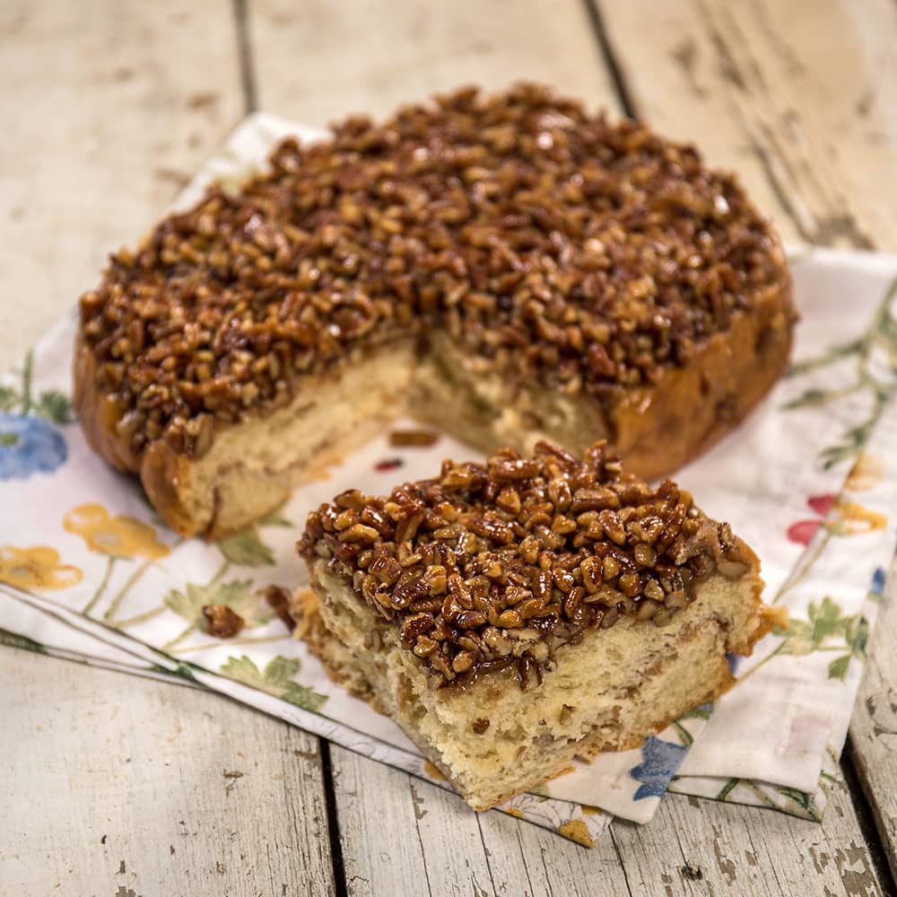Close-up of a freshly baked pecan roll slice topped with chopped pecans and caramel glaze, handcrafted and made in USA, presented on a floral cloth in a rustic kitchen setting.