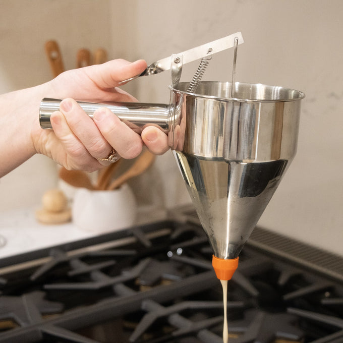 Handheld stainless steel batter dispenser with thumb lever and orange silicone tip releasing batter, featured in Kitchen & Food, Cookware, and Baking Supplies collections.