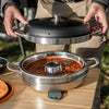 Person lifting the black domed lid on a stainless steel camping oven filled with simmering stew, placed on a wooden table outdoors, ideal for Farm, Garden & Tool and Camping collections