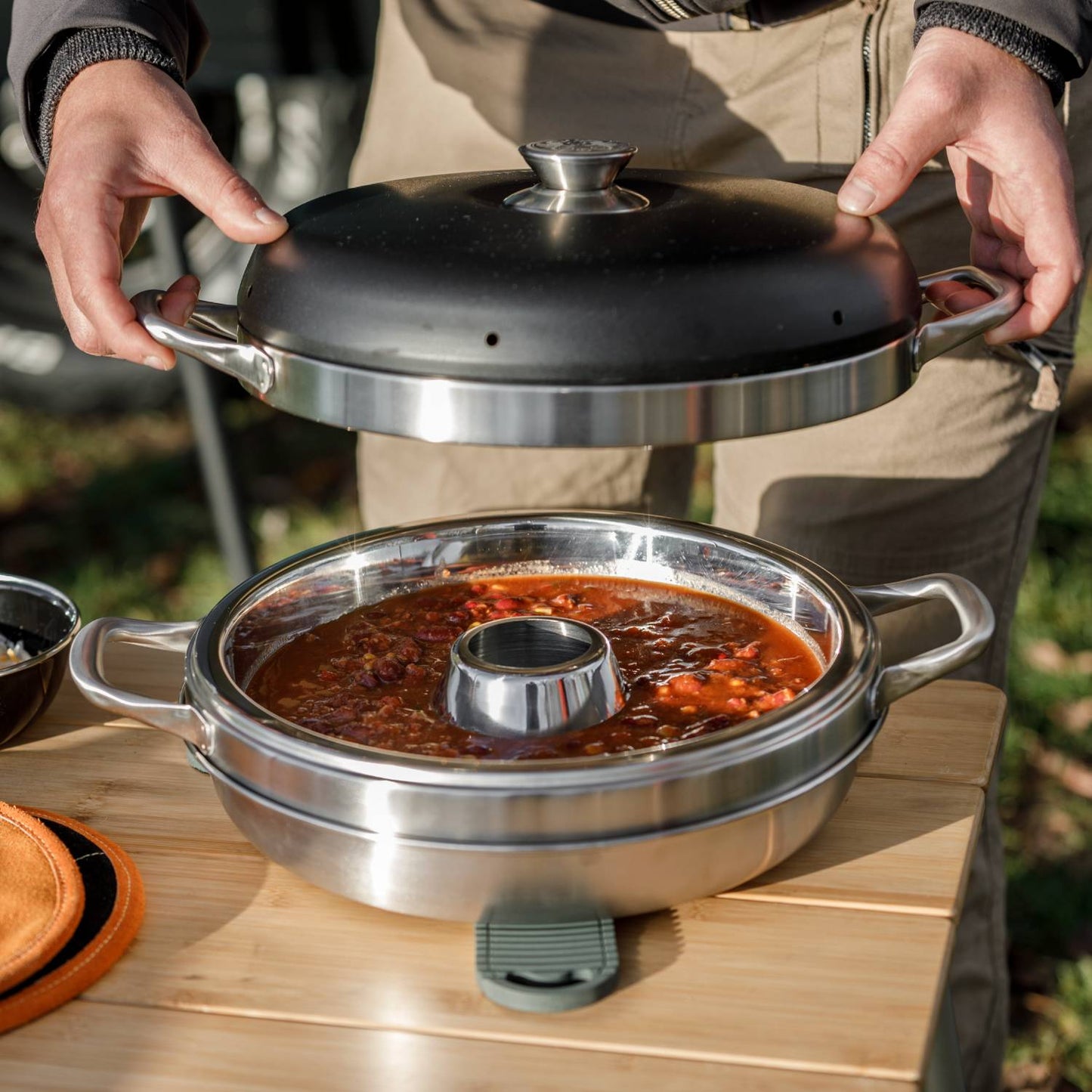 Person lifting the black domed lid on a stainless steel camping oven filled with simmering stew, placed on a wooden table outdoors, ideal for Farm, Garden & Tool and Camping collections