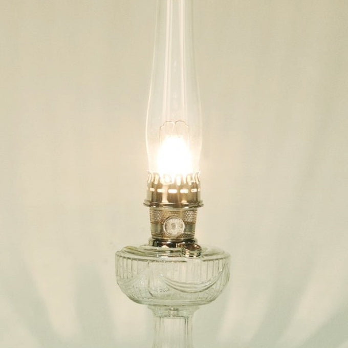 Lit clear glass oil lamp with a tall chimney and metal burner, emitting a warm glow, featured in Lamps & Lighting, Parts and Accessories, Wicks and Mantles, Lamp Burners and Collars, Best Sellers, and Promo Eligible collections.