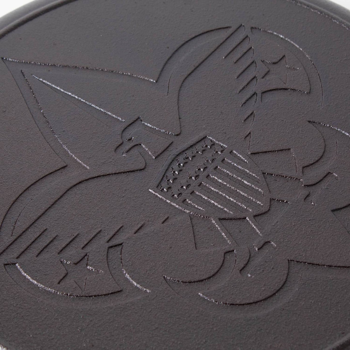 Close-up of cast iron skillet surface featuring embossed Boy Scout emblem with eagle, shield, and fleur-de-lis design, highlighting durable seasoning and USA craftsmanship.