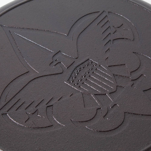Close-up of cast iron skillet surface featuring embossed Boy Scout emblem with eagle, shield, and fleur-de-lis design, highlighting durable seasoning and USA craftsmanship.