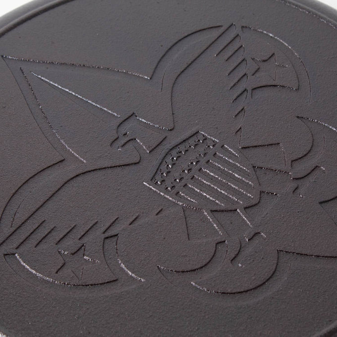 Close-up of cast iron skillet surface featuring embossed Boy Scout emblem with eagle, shield, and fleur-de-lis design, highlighting durable seasoning and USA craftsmanship.