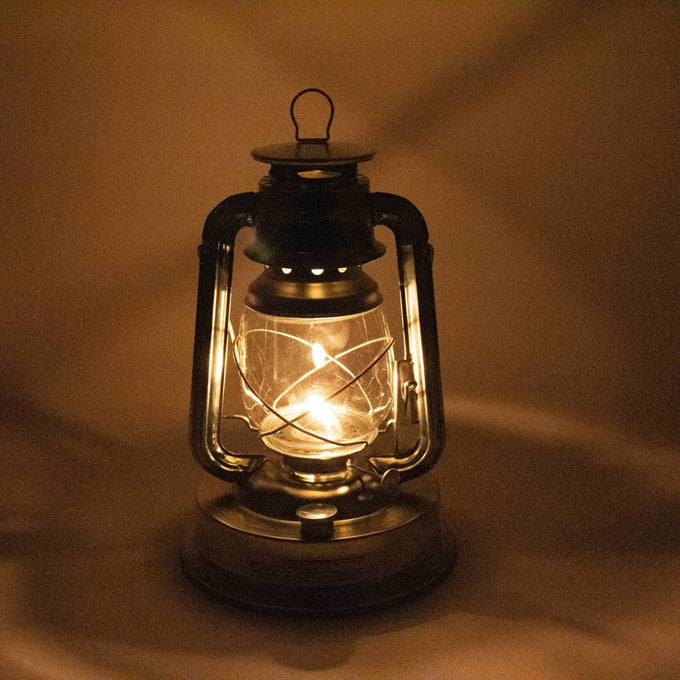 Lit handcrafted metal oil lantern casting soft warm light, ideal for outdoor use in camping, farm, garden, pest control, and emergency supplies collections.
