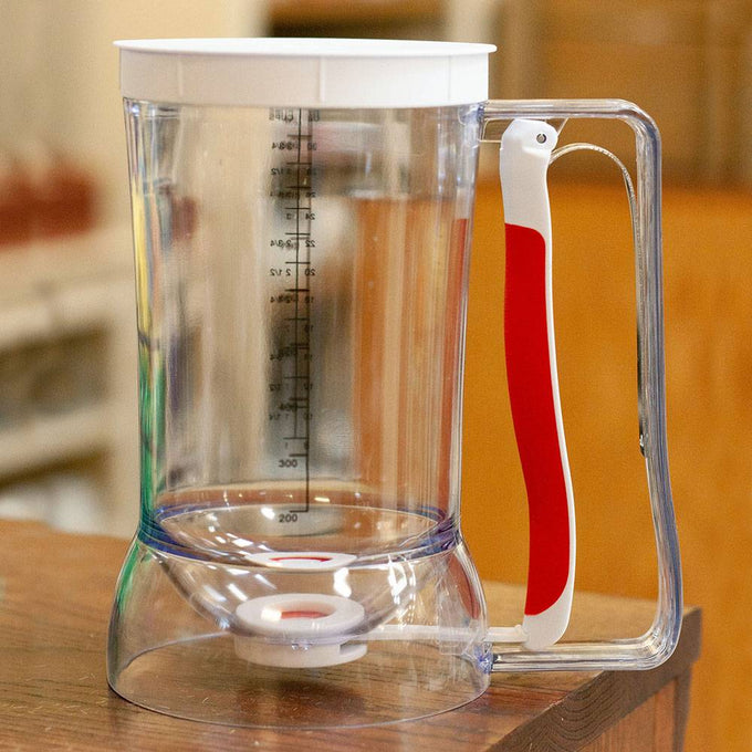 Clear plastic batter dispenser with ergonomic white and red soft-grip handle, featuring measurement markings for precise pouring, ideal for Kitchen & Food, Baking Helpers, and Promo Eligible collections.