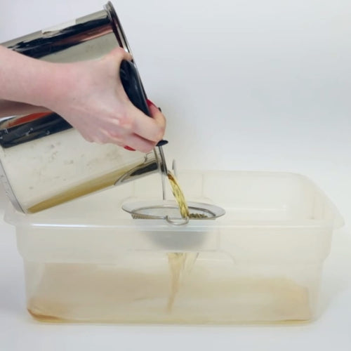 Hand pouring herbal soak solution from a stainless steel container through a fine mesh strainer into a clear plastic basin, showcasing natural health and wellness preparation in USA Made collections.