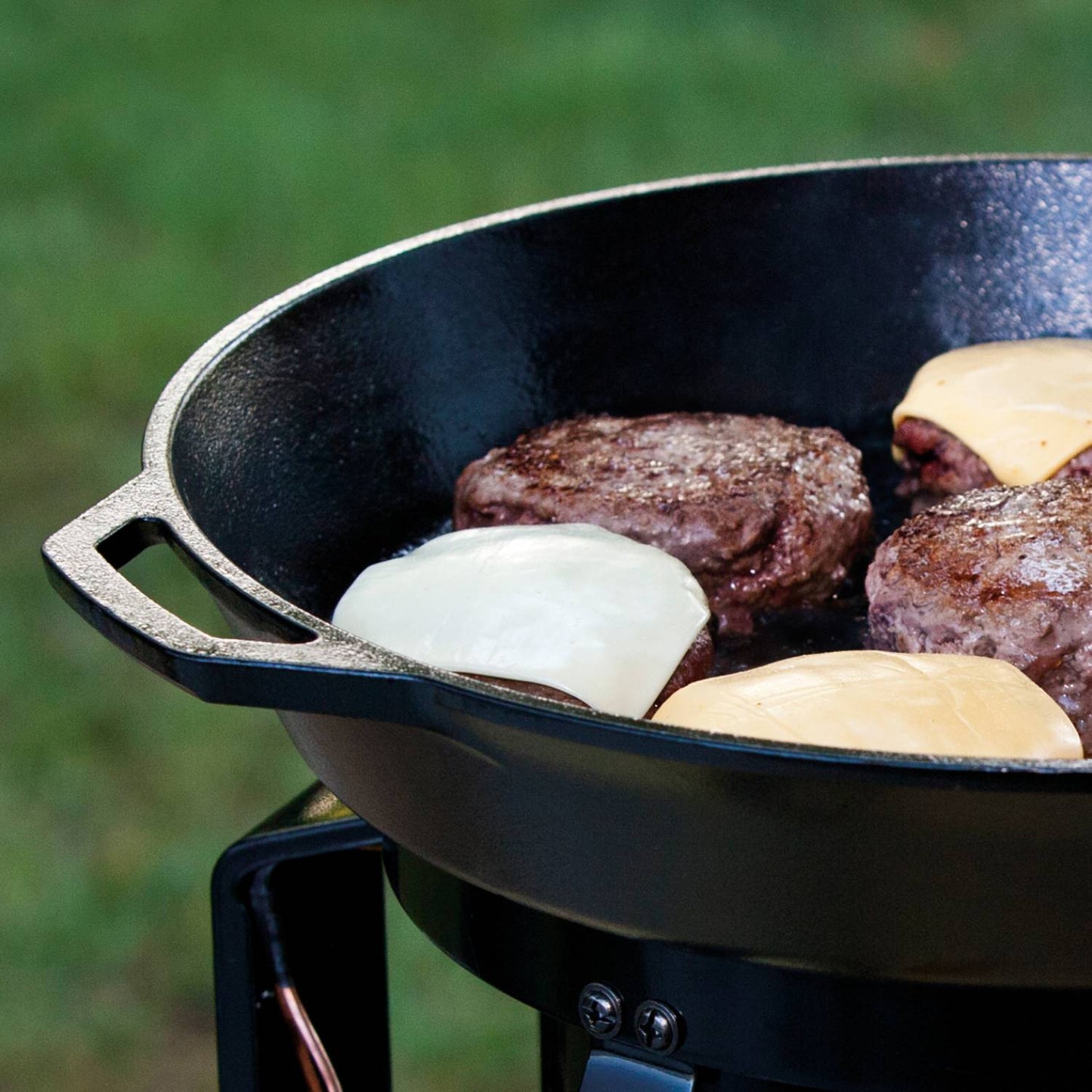 Close-up of a large cast iron skillet cooking four cheeseburgers outdoors on a portable stove, showcasing durable cookware from Kitchen & Food and Cookware collections, USA made.