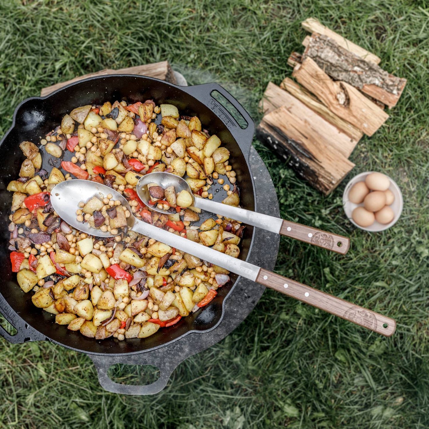 Two long-handled stainless steel spoons with walnut handles resting in a cast iron skillet of cooked potatoes, chickpeas, and vegetables, on grass with firewood and a bowl of eggs nearby, crafted for Farm, Garden & Tool, Kitchen & Food, Camping, Storage and Serving, Best Sellers, Promo Eligible collections.