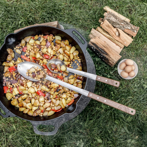 Two long-handled stainless steel spoons with walnut handles resting in a cast iron skillet of cooked potatoes, chickpeas, and vegetables, on grass with firewood and a bowl of eggs nearby, crafted for Farm, Garden & Tool, Kitchen & Food, Camping, Storage and Serving, Best Sellers, Promo Eligible collections.