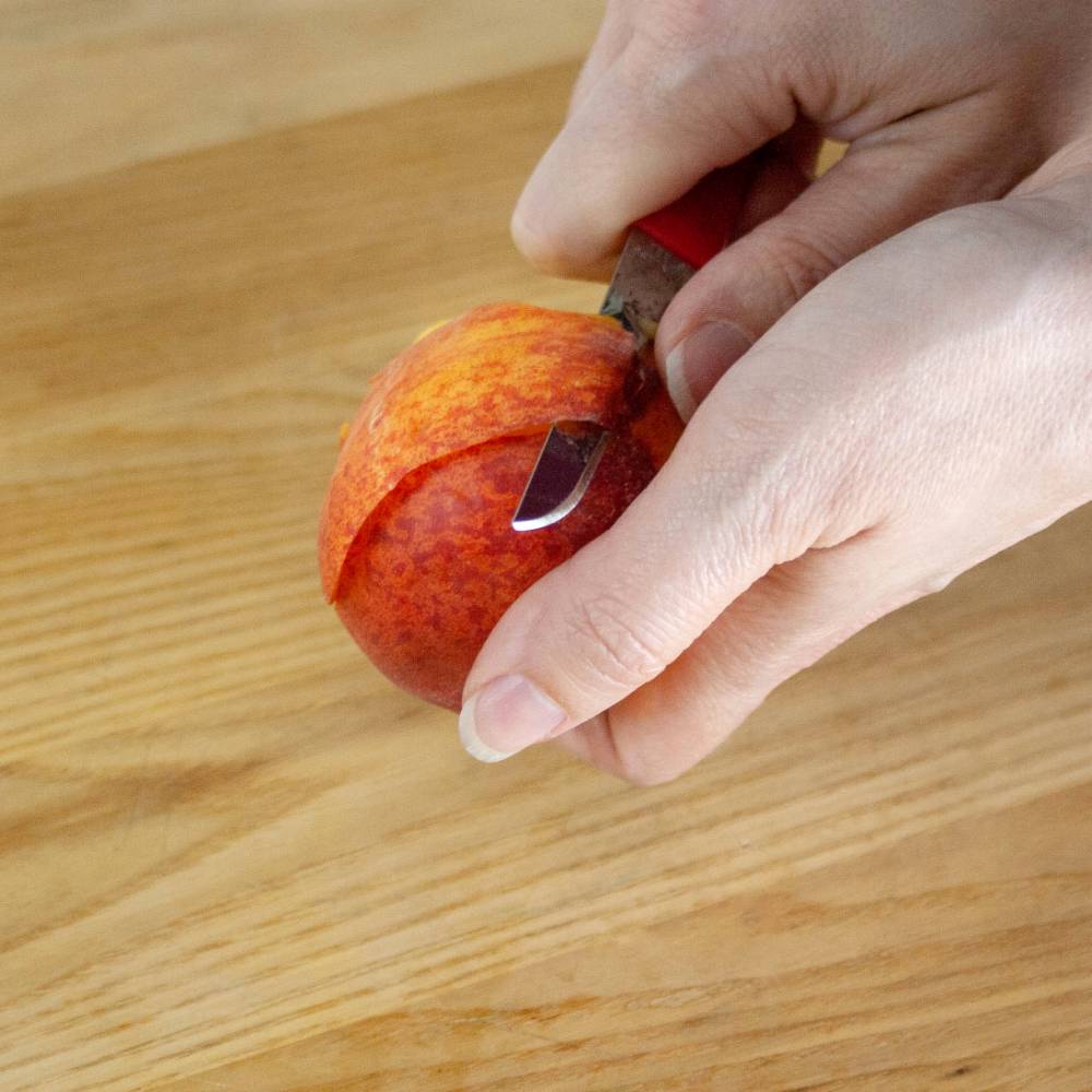 Hand using a handcrafted flexible paring knife with a red handle to peel a small orange fruit on a wooden surface, part of Farm, Garden & Tool and Kitchen & Food collections.