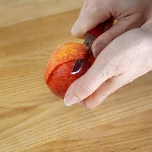 Hand using a handcrafted flexible paring knife with a red handle to peel a small orange fruit on a wooden surface, part of Farm, Garden & Tool and Kitchen & Food collections.