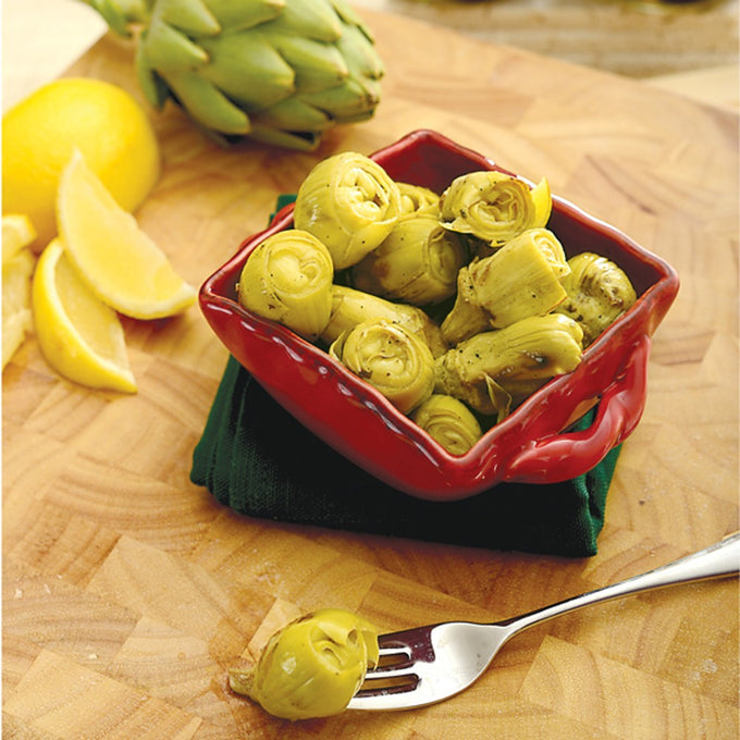 Red ceramic dish filled with fire-grilled artichoke hearts on a wooden surface, accompanied by lemon wedges and fresh artichoke, featured in Kitchen & Food and Pantry collections.