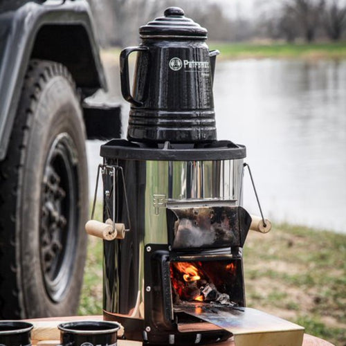 Black enameled steel percolator on a portable wood-burning stove near a riverside, ideal for brewing aromatic coffee or tea, featured in Farm, Garden & Tool, Camping, Kitchen & Food, and Brewing Coffee and Tea collections.