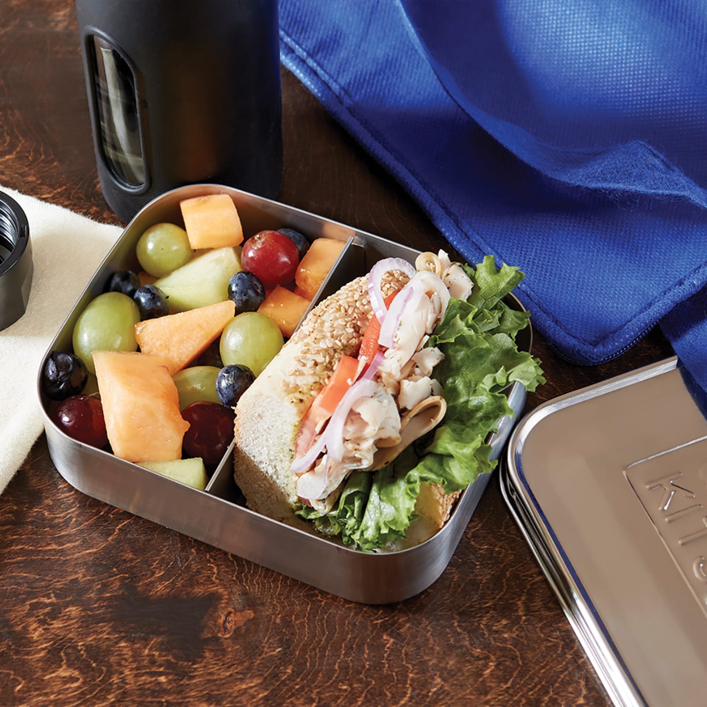 Open stainless steel container featuring two compartments, one holding a fresh sandwich with lettuce and sliced deli meat, the other filled with mixed fruit, ideal for Kitchen & Food, Storage and Serving, Best Sellers, and Promo Eligible collections.