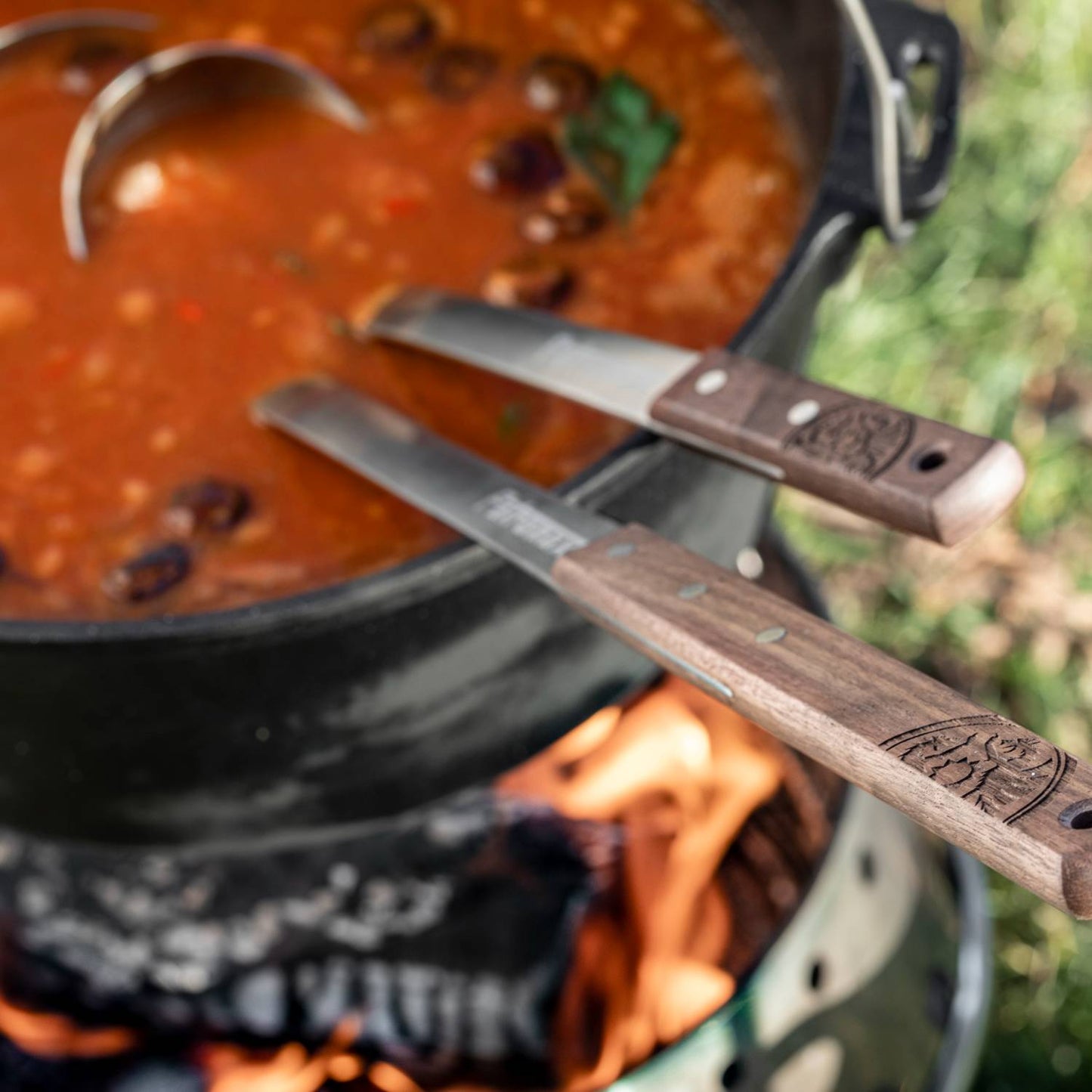 Generously sized stainless steel soup ladle with engraved wooden handle resting on a cast iron pot of simmering soup over an open flame, ideal for Farm, Garden & Tool, Kitchen & Food, and Camping collections.