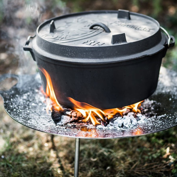 Sturdy cast iron Dutch oven with legs cooking over an open flame on a metal stand, featuring a detailed embossed lid and side handles, ideal for Kitchen & Food, Cookware, Cast Iron, Best Sellers, Promo Eligible collections.