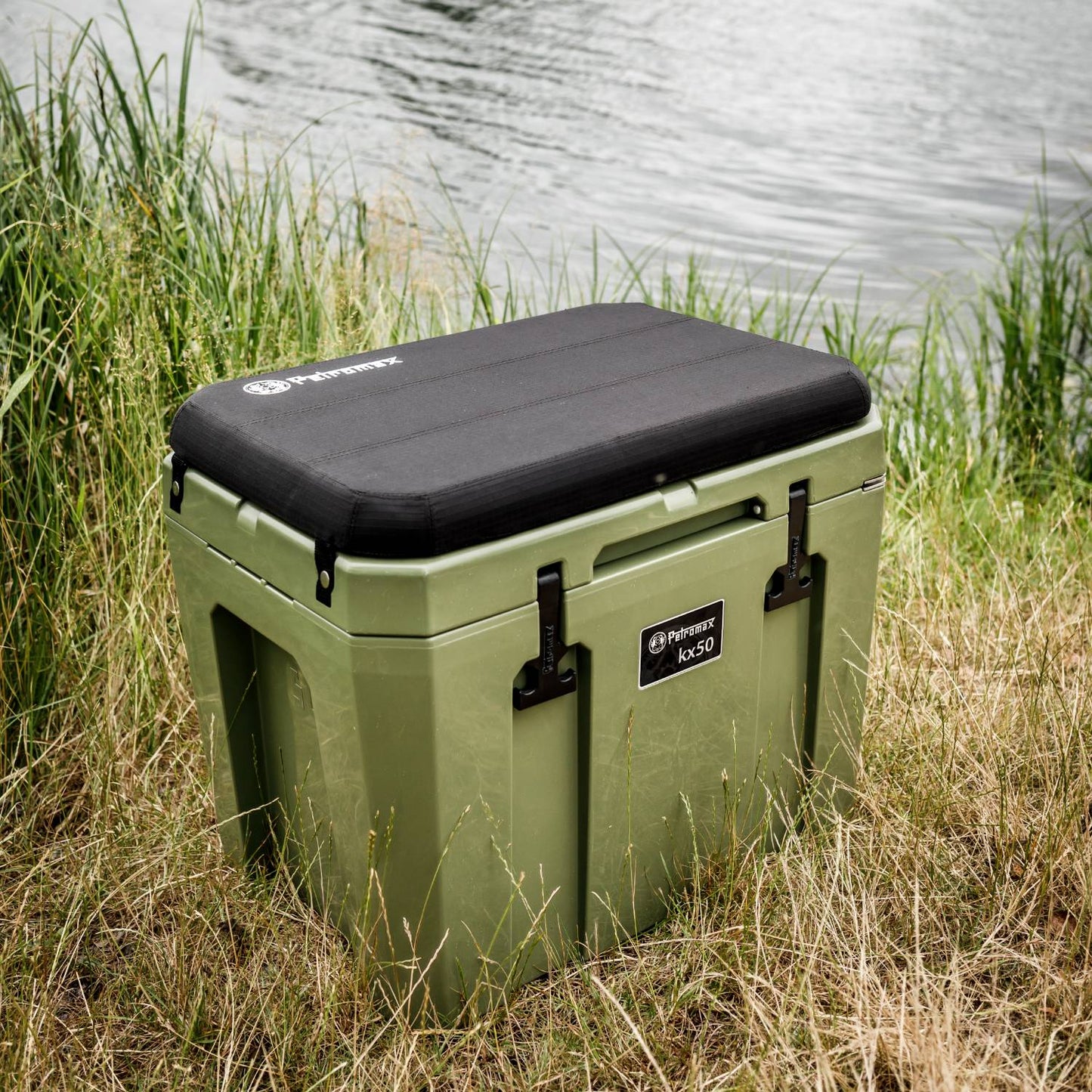 Olive green cooler with black cushioned lid and sturdy latches, set on grassy bank beside a calm lake, designed for reliable outdoor use in Farm, Garden & Tool, Camping, and Best Sellers collections.