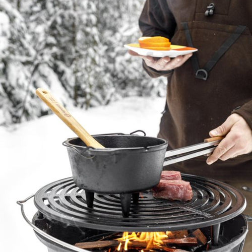 Three-legged cast iron Dutch oven simmering on a round outdoor grill over a wood fire with raw meat nearby, a wooden stirrer inside, suited for Kitchen & Food, Cookware, Cast Iron collections.