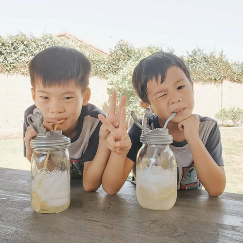 Two young boys outdoors sitting at a wooden table enjoying beverages in glass jars with sturdy gray pour lids and carry loops, perfect for Kitchen Gadgets, Kitchen Accessories, USA Made, and Best Sellers collections.