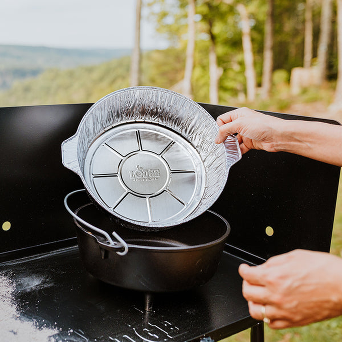 Person placing a heavy gauge aluminum foil liner into a black cast iron Dutch oven on an outdoor camp stove, showcasing cookware and kitchen gadgets for easy clean-up during camping trips.