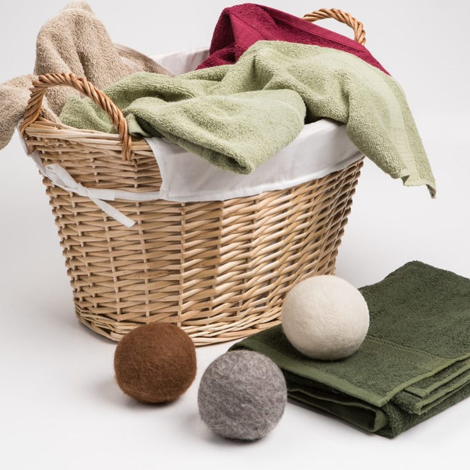 Three reusable wool dryer balls in natural brown, gray, and cream colors placed near a wicker laundry basket with folded green and beige towels, featured in USA Made and Home & Decor collections.