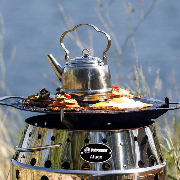 Oiled steel grilling plate with multiple temperature zones cooking eggs, vegetables, and a kettle on a portable stainless steel stove in outdoor camping and grilling setting.