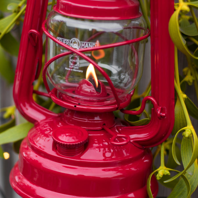 Handcrafted bright red galvanized oil lantern with durable thermal heat-resistant SUPRAX globe, gently glowing amidst green foliage, ideal for Camping, Lamps & Lighting, and Emergency Supplies collections.