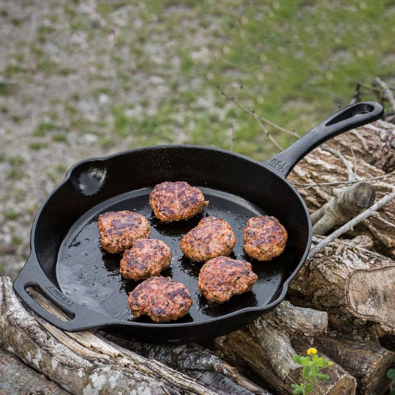 Cast iron fire skillet with long handle cooking seven browned meat patties outdoors on stacked firewood, ideal for Farm, Garden & Tool, Camping, Kitchen & Food, Cookware, Cast Iron, Best Sellers, and Promo Eligible collections.