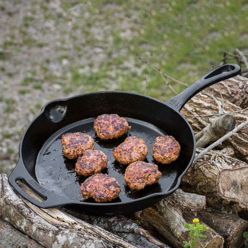 Cast iron fire skillet with long handle cooking seven browned meat patties outdoors on stacked firewood, ideal for Farm, Garden & Tool, Camping, Kitchen & Food, Cookware, Cast Iron, Best Sellers, and Promo Eligible collections.