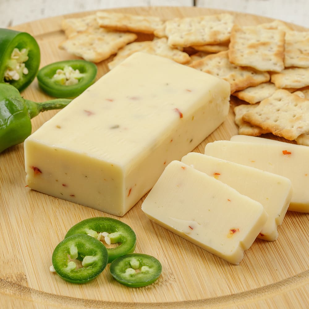 Handcrafted shelf-stable cheese block with visible chili flakes, sliced and served on a wooden board with crackers and fresh jalapeño slices, ideal for Kitchen & Food, Snacks and Sweets, Pantry, USA Made, and Best Sellers collections.