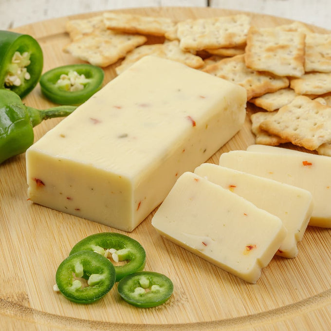 Handcrafted shelf-stable cheese block with visible chili flakes, sliced and served on a wooden board with crackers and fresh jalapeño slices, ideal for Kitchen & Food, Snacks and Sweets, Pantry, USA Made, and Best Sellers collections.