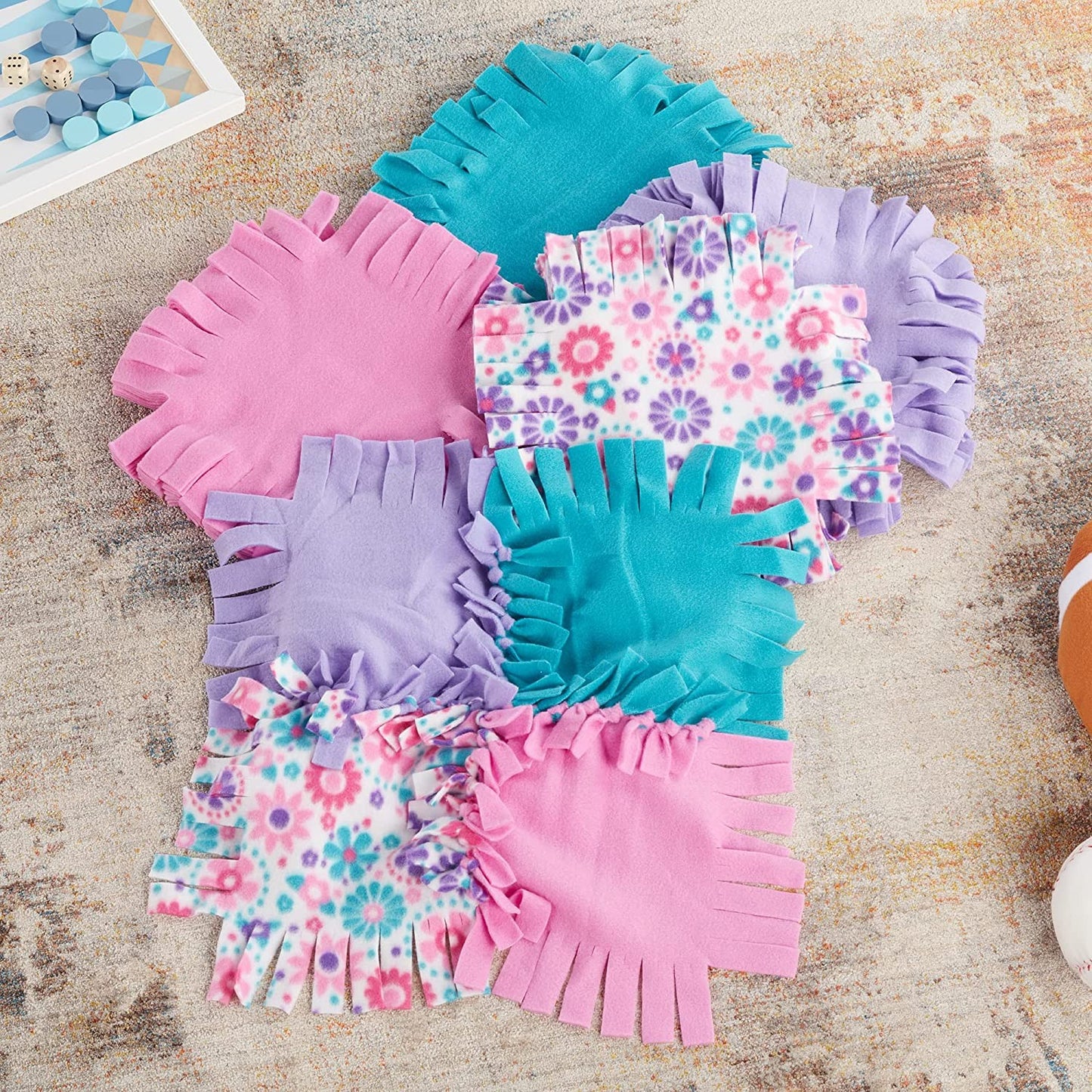 Colorful handcrafted fleece squares in pink, purple, teal, and floral patterns with fringe edges arranged on a textured surface, ideal for children’s arts and crafts and toys collections.
