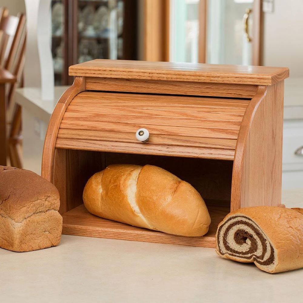 Light honey-brown oak roll-top bread box with smooth finish and white knob, handcrafted in USA, Amish made, shown with assorted fresh bread, ideal for Kitchen & Food and Storage collections.