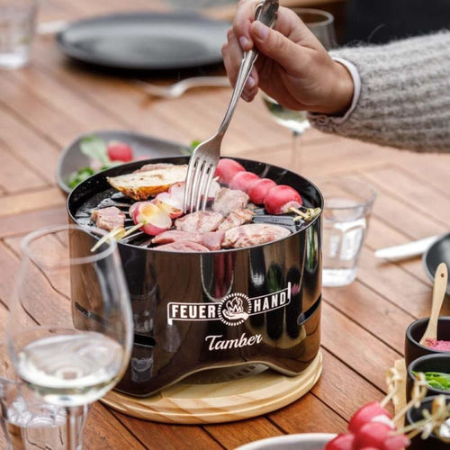 Glossy black mini charcoal grill with food cooking on a circular grate, set on a wooden trivet amid drinks and dishes, perfect for cozy outdoor grilling in Farm, Garden & Tool and Camping collections.
