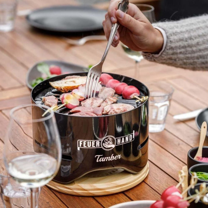 Glossy black mini charcoal grill with food cooking on a circular grate, set on a wooden trivet amid drinks and dishes, perfect for cozy outdoor grilling in Farm, Garden & Tool and Camping collections.