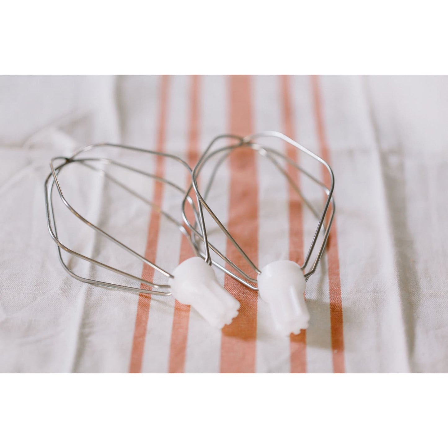 Two handcrafted batter whisk attachments with polished metal wires and white plastic connectors, resting on a striped kitchen towel, ideal for Kitchen & Food and Baking Supplies collections.