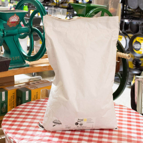 Large white paper sack of organic whole amaranth grain packaged for pantry and emergency supplies, displayed in a kitchen setting with grain mills and food prep tools nearby.