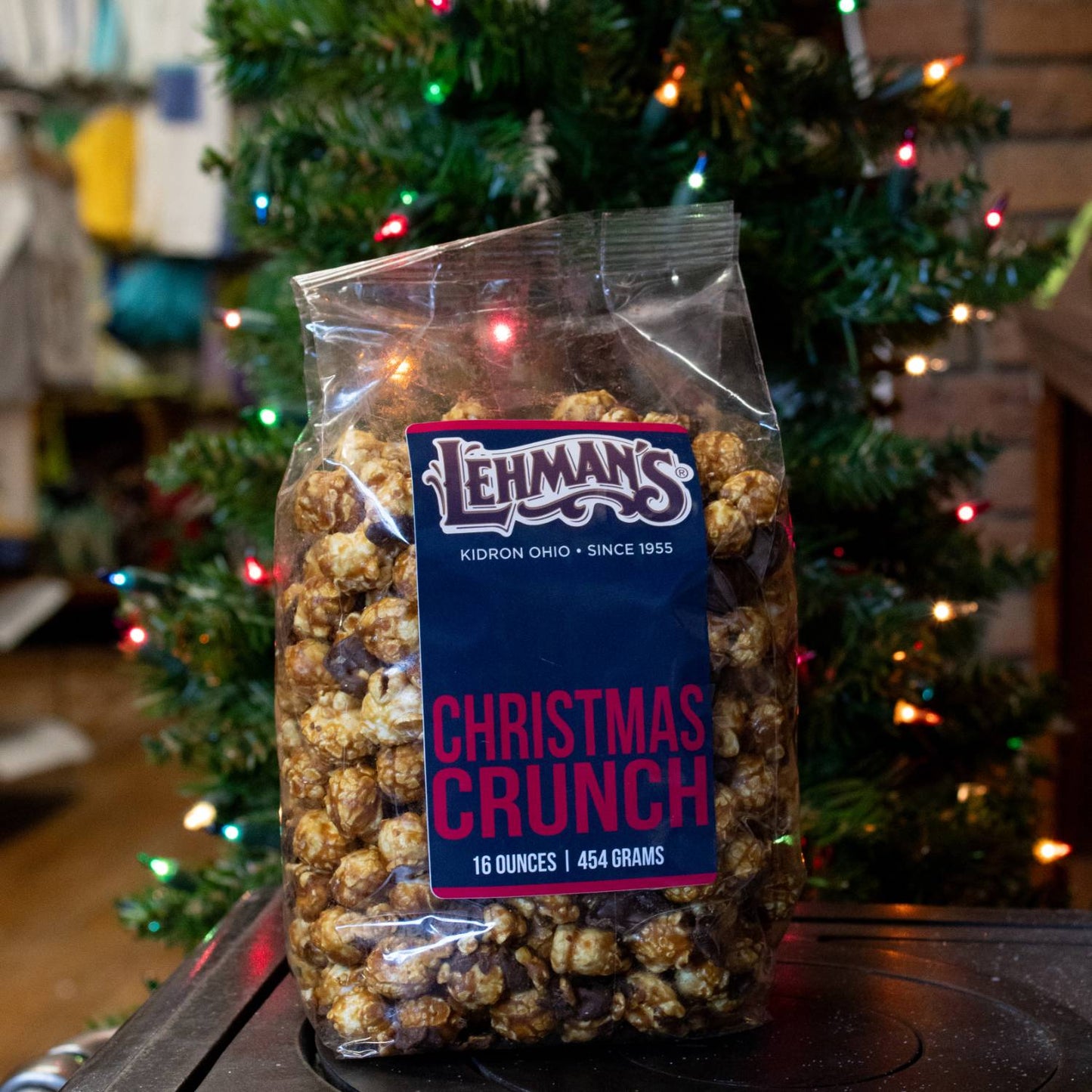 Clear bag of handcrafted caramel popcorn mixed with festive holiday ingredients and labeled Christmas Crunch, displayed in a cozy home setting with a decorated Christmas tree, featured in Kitchen & Food, Snacks and Sweets, Popcorn, and Best Sellers collections.