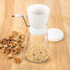 Compact nut chopper with a white plastic hopper, glass jar base filled with chopped nuts, a metal crank handle, and a white lid, perfect for kitchen food prep and choppers and cutters collections.