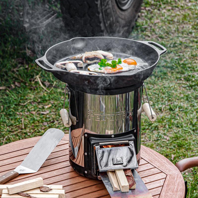 Handcrafted cast iron skillet with two handles cooking eggs and mushrooms on a compact outdoor fire stove, perfect for farm, camping, kitchen, cookware, and cast iron collections.