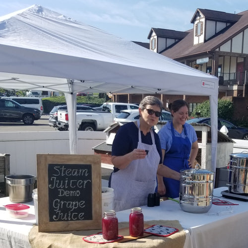 Two women wearing aprons demonstrating a stainless steel steam juicer under a white canopy tent at an outdoor event, with jars of grape juice on the table in front, featured in Kitchen & Food, Juicers and Steamers, Best Sellers, and Promo Eligible collections.
