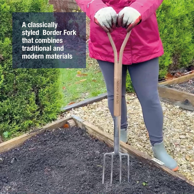 Person wearing gloves and pink jacket holding a traditional-style border fork with a stainless steel head and wooden handle, positioned over garden soil, suitable for Farm, Garden & Tool, Digging and Cultivating, Garden Accessories, Lawncare, Best Sellers, and Promo Eligible collections.