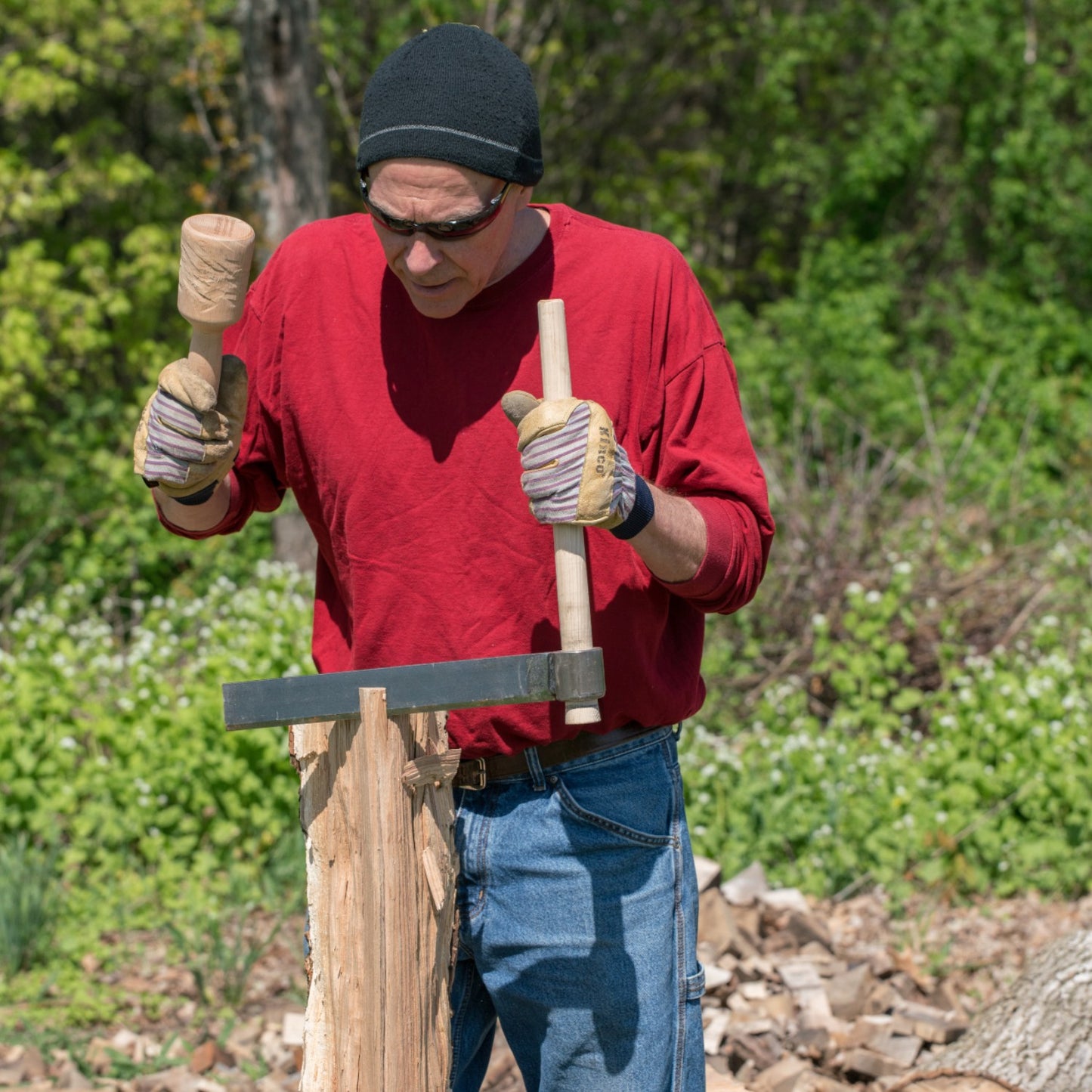 Man wearing gloves and sunglasses using a handcrafted wood splitting froe with a wooden mallet to split a log outdoors, set against a forested background in a farm and garden tool setting.